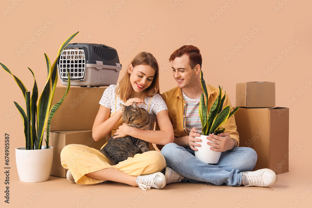 Young couple with cute cat and moving boxes on beige background