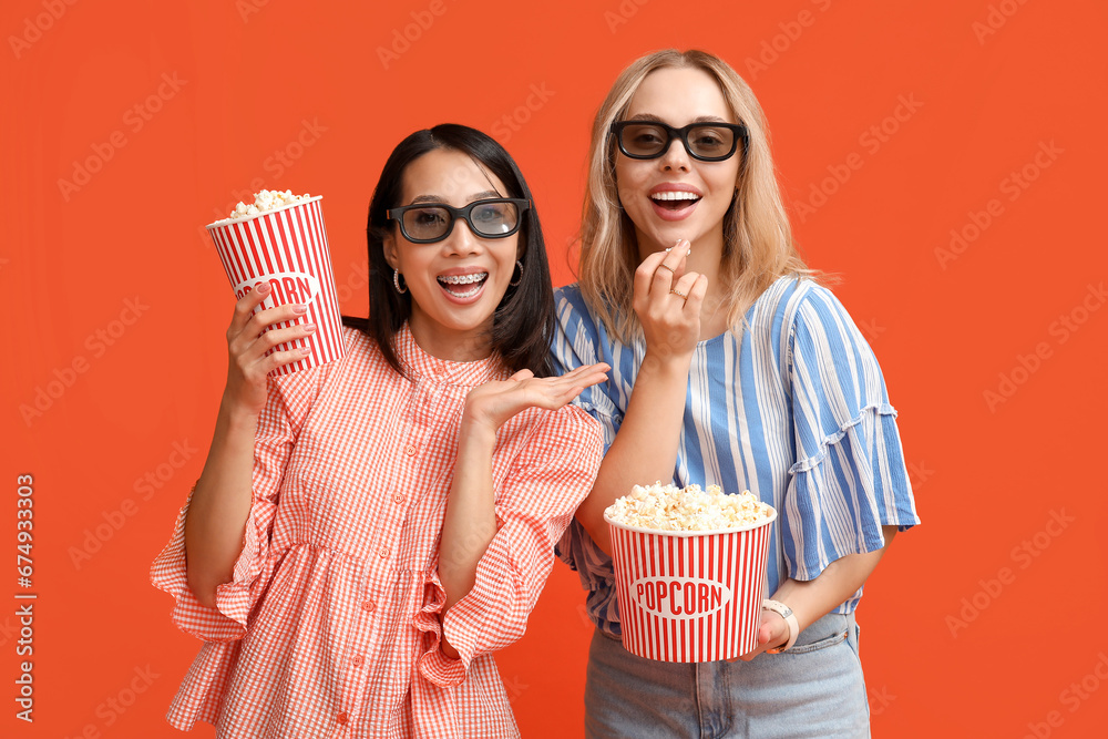 Beautiful young women in 3D glasses with buckets of popcorn on orange background