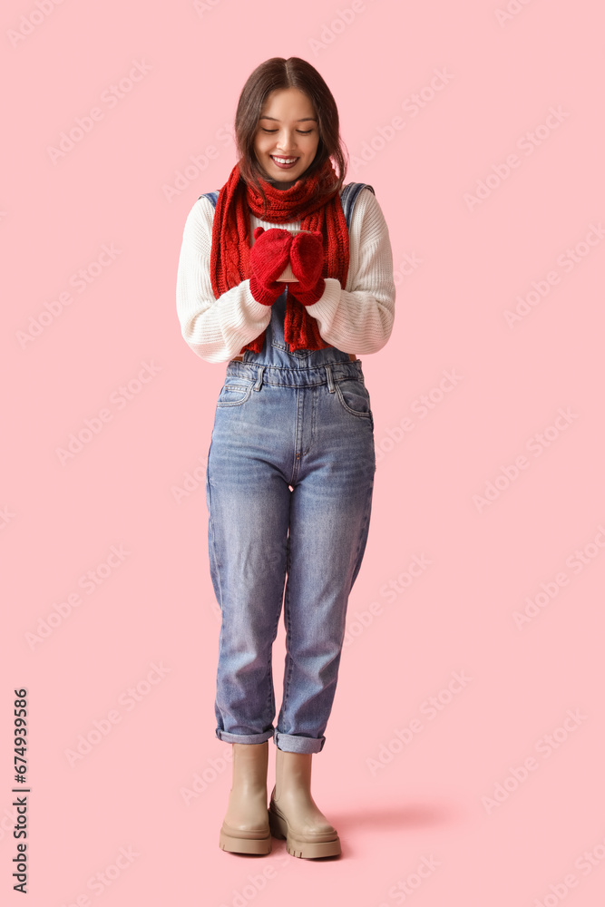 Beautiful young Asian woman in warm scarf with cup of tea on pink background