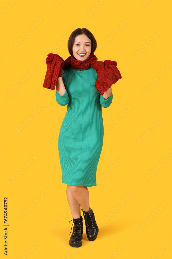 Beautiful young Asian woman in warm scarf and gloves on yellow background