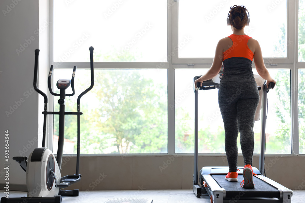 Beautiful young woman running on treadmill in gym while listening to music, back view