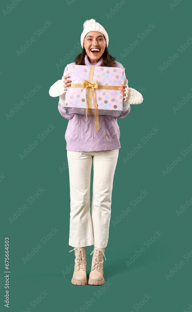 Young woman in winter clothes with gift on green background