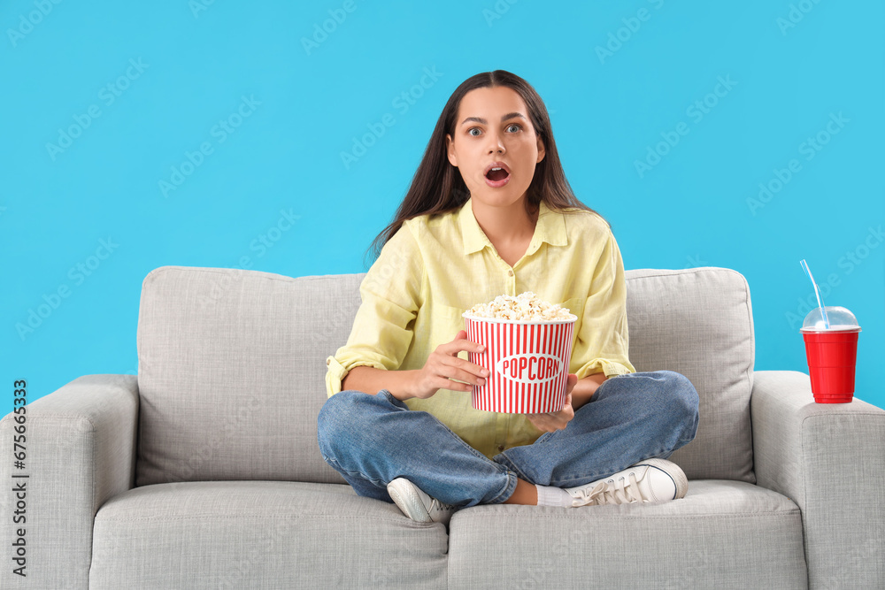 Shocked young woman with popcorn watching movie on sofa against blue background