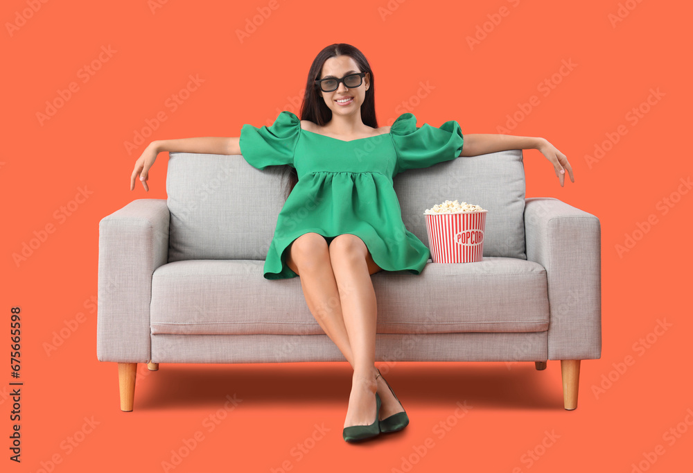 Young woman in 3D glasses with popcorn watching movie on sofa against orange background