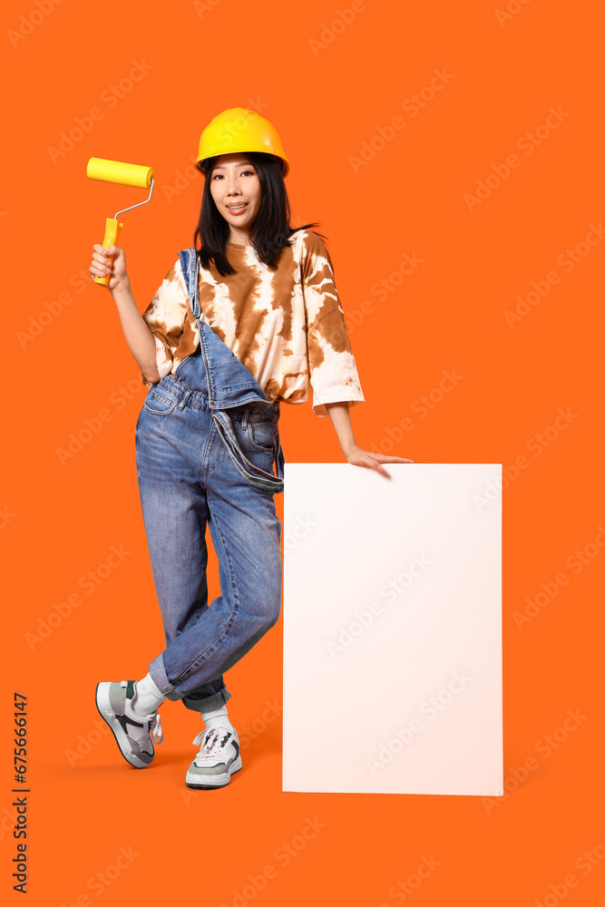 Young Asian woman with paint roller and blank poster on orange background