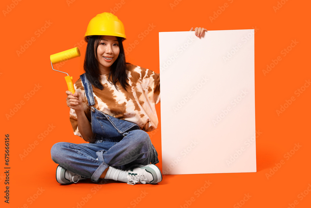 Young Asian woman with paint roller and blank poster on orange background