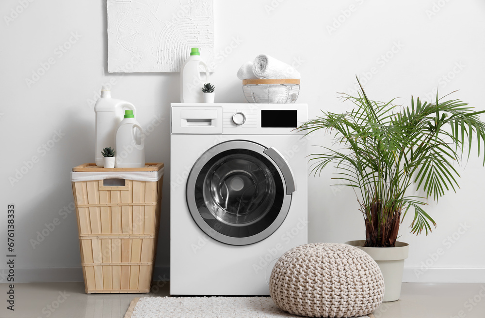 Washing machine with bottles of detergent, laundry basket and pouf near white wall