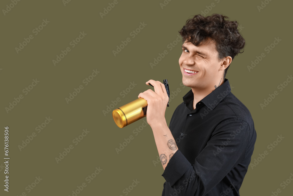 Handsome young man with bottle of hair spray winking on green background