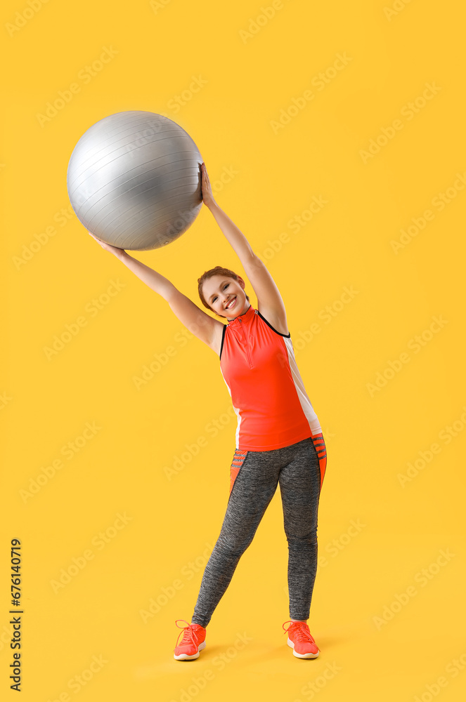 Beautiful young woman in sportswear exercising with fitness ball on yellow background