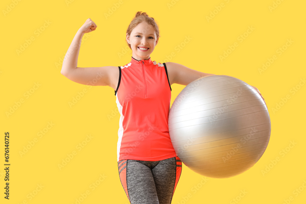 Beautiful young woman with fitness ball flexing her muscles on yellow background