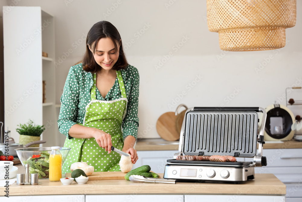 Beautiful young woman cutting mushrooms near modern electric grill with sausages at kitchen