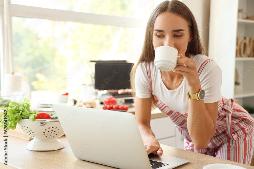 Beautiful young woman using laptop while cooking delicious sausages on modern electric grill in kitchen