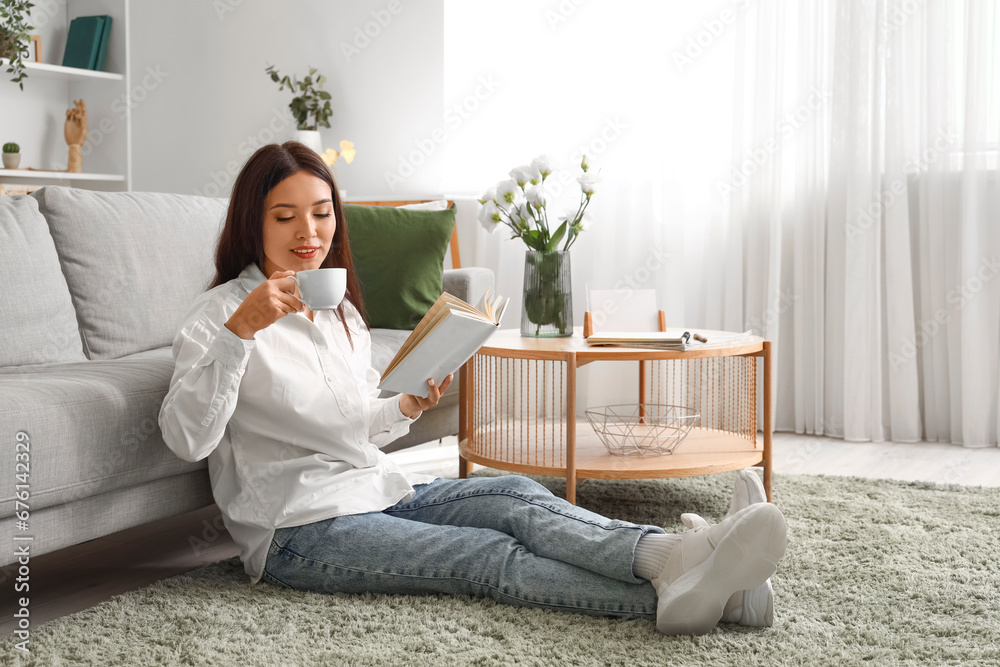 Beautiful young Asian woman with cup of tea reading book in living room