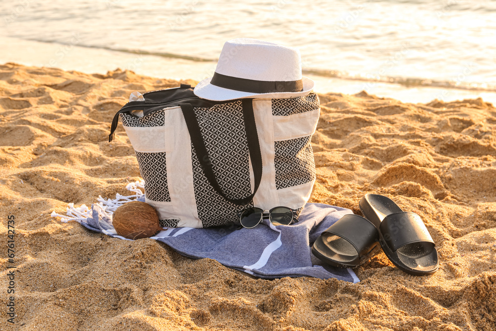 Stylish beach bag, hat, sunglasses, coconut, flip-flops and towel on sand near sea