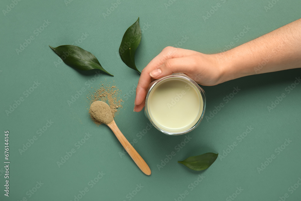 Female hand with cup of fresh matcha tea, powder and plant leaves on green background