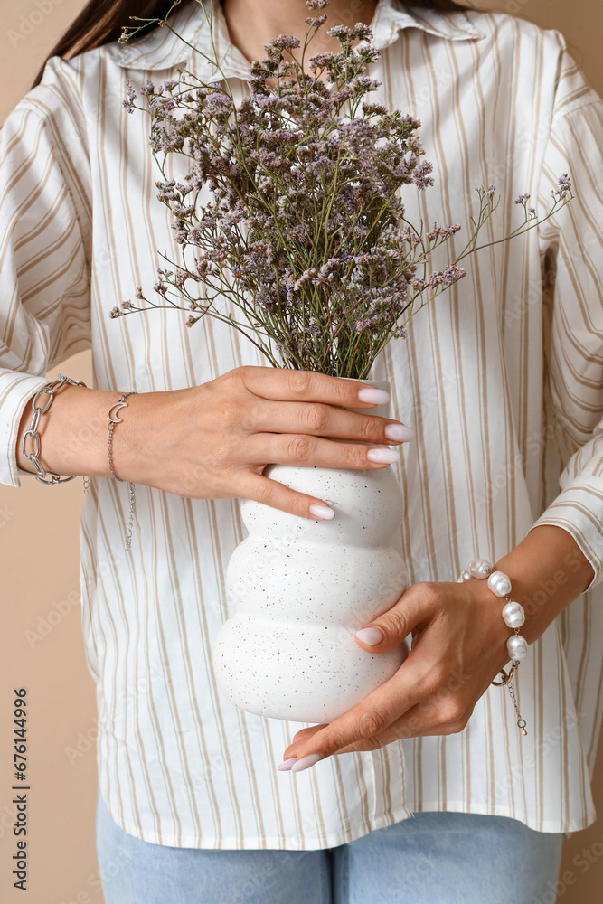 Stylish young woman with bracelets and flowers in vase on beige background, closeup