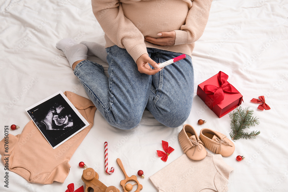 Pregnant woman with test, baby accessories and Christmas decorations on bed