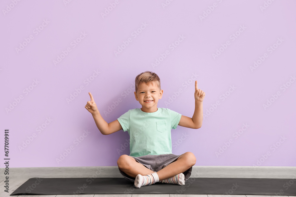 Little boy sitting on yoga mat and pointing at something against lilac wall