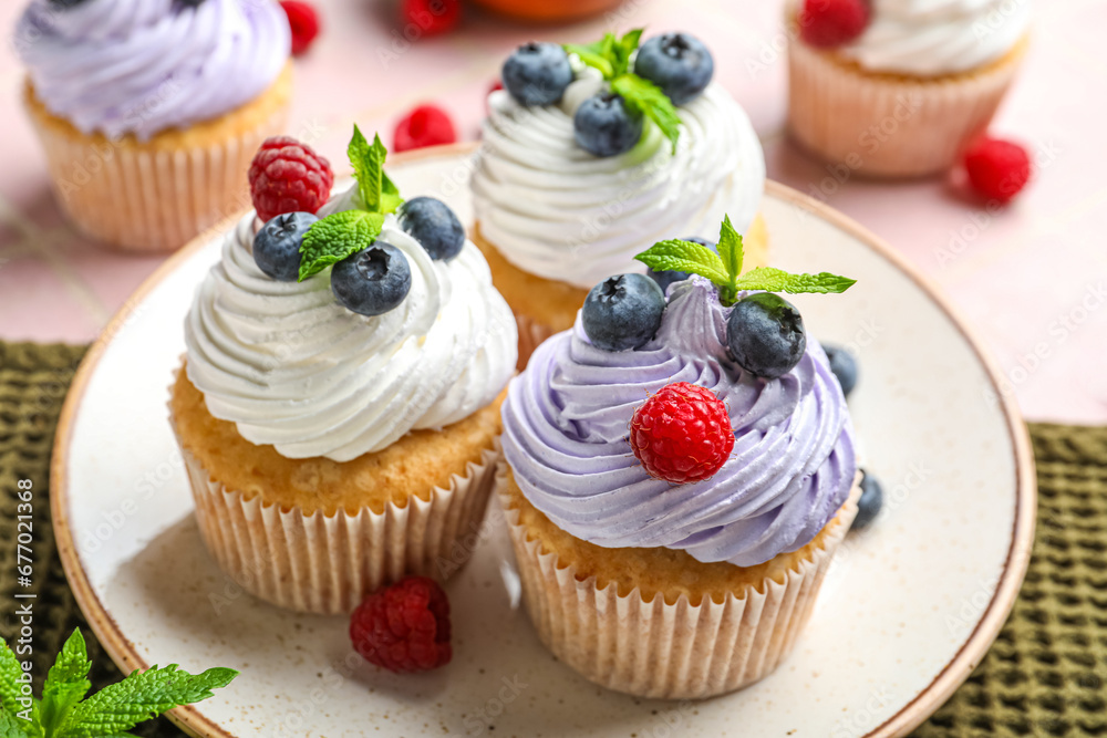 Plate of delicious cupcakes with blueberries, raspberries and mint on pink tile background