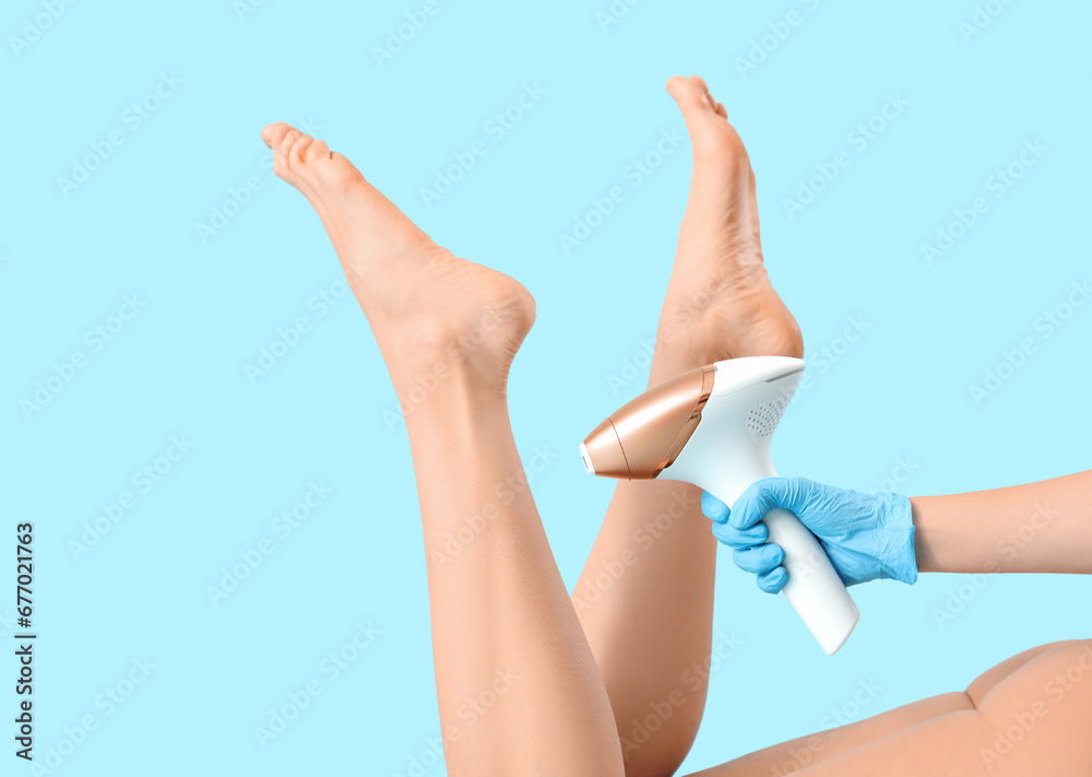 Beautiful young woman and hand epilating her legs with modern photoepilator on blue background