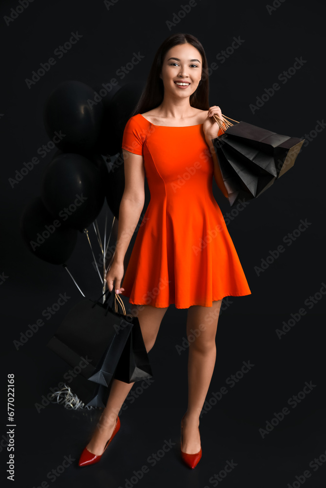 Beautiful young Asian woman with shopping bags and balloons on dark background. Black Friday sale