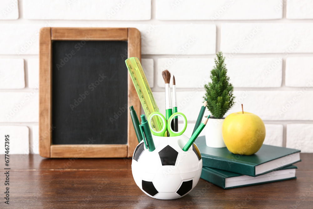 Holder in shape of soccer ball with different stationery, tasty apple and chalkboard on wooden desk, closeup