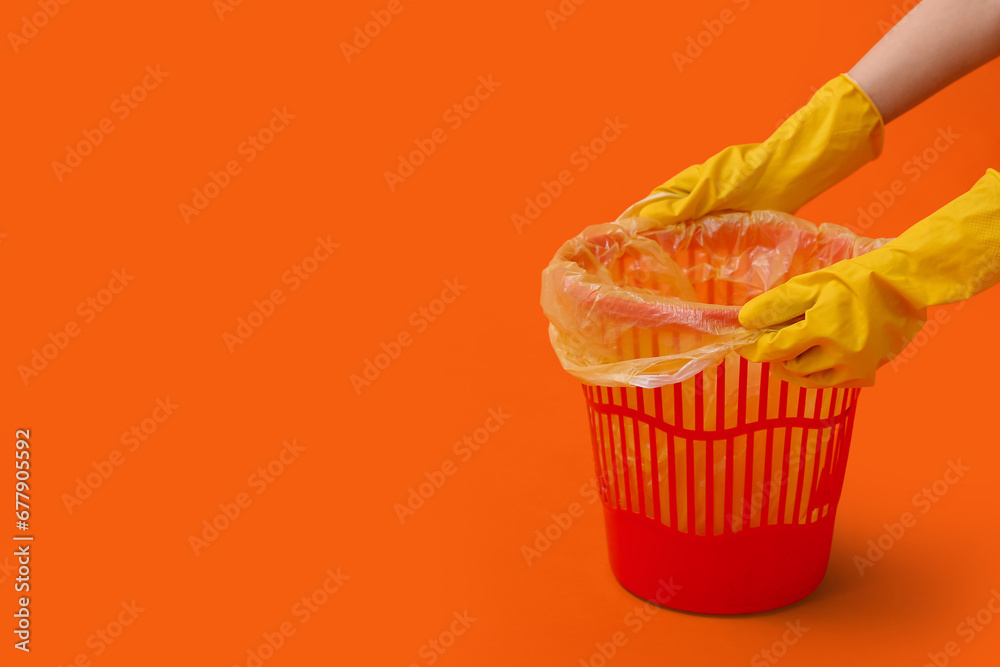 Female hands in rubber gloves placing garbage bag into trash bin on orange background