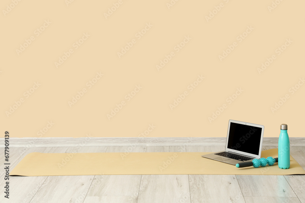 Laptop with bottle of water and massage roller on fitness mat near beige wall in room