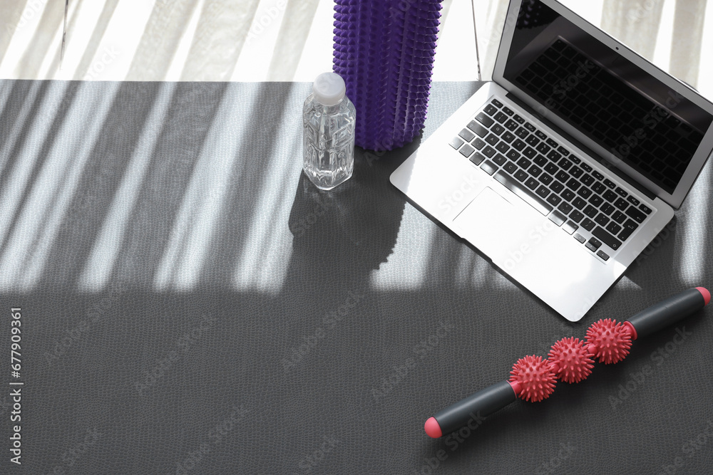 Laptop with bottle of water and rollers on fitness mat in gym