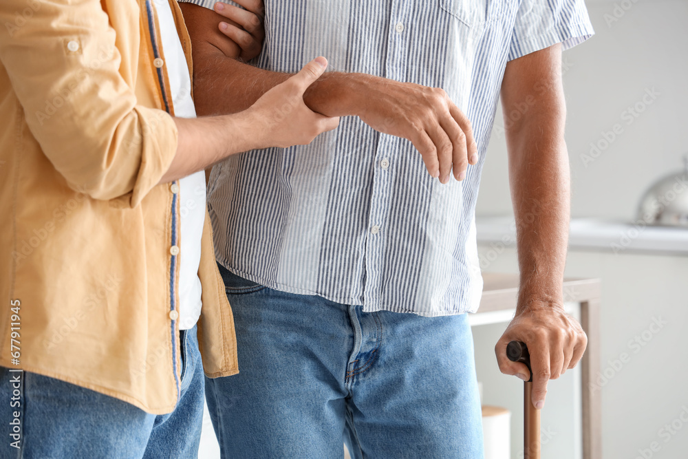 Young man helping his father with stick to walk in kitchen, closeup