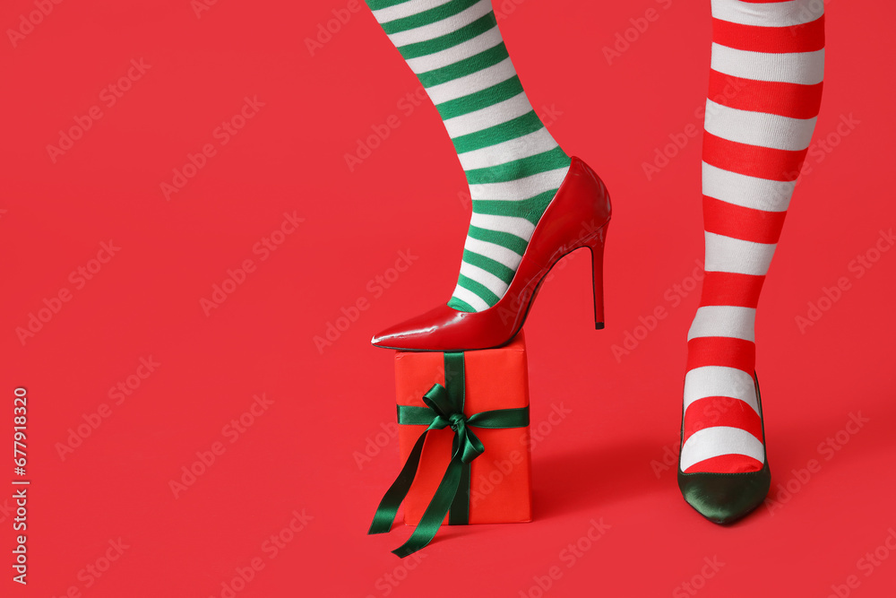 Legs of young woman in striped stockings with Christmas gift box on red background