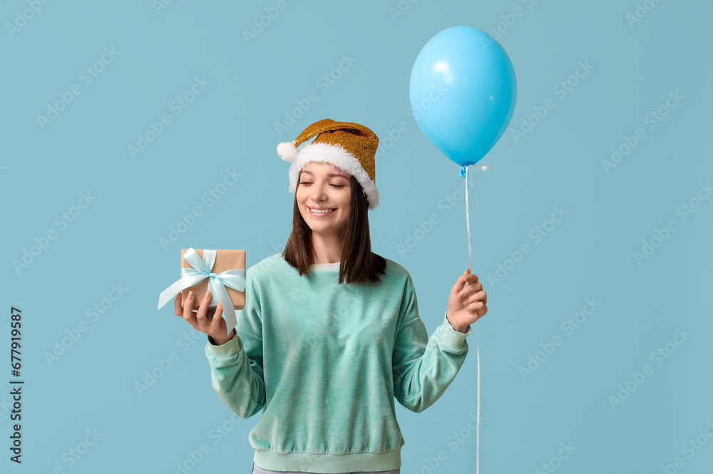 Beautiful young woman in Santa hat with balloon and gift box on blue background