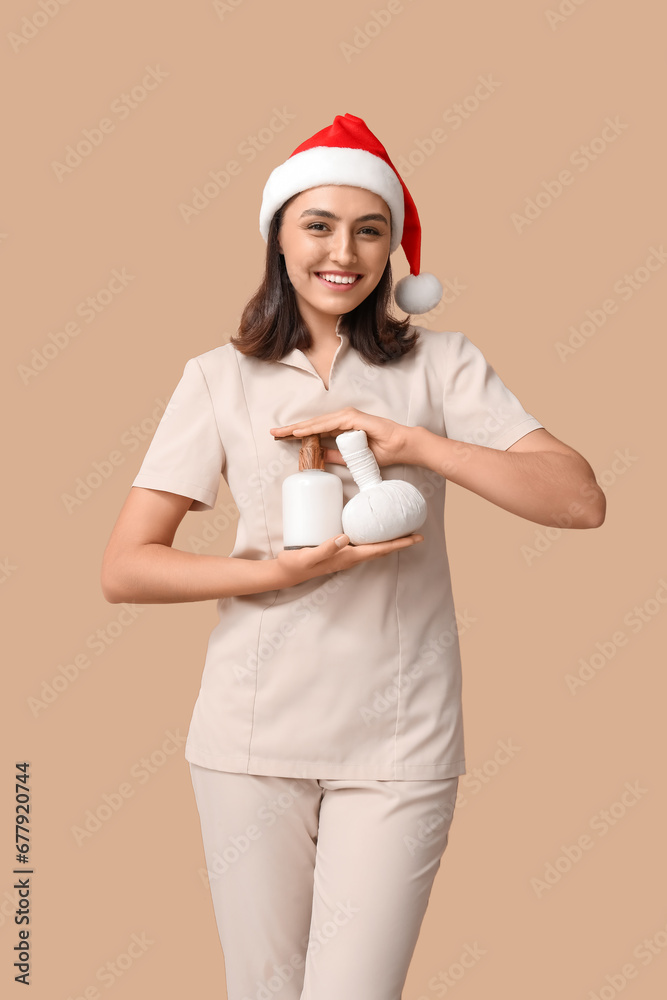 Beautiful female masseur in Santa hat with massage herbal ball and lotion on brown background