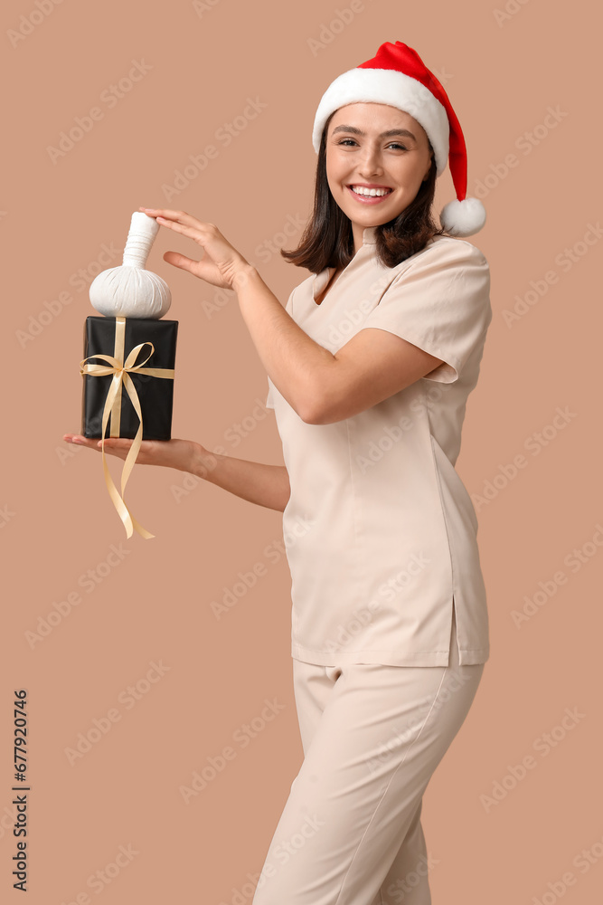 Beautiful female masseur in Santa hat with gift box and massage herbal ball on brown background