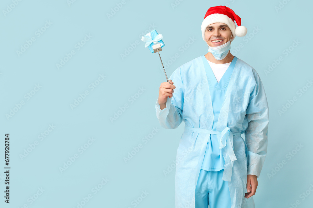 Male surgeon in Santa hat with gift box and forceps on blue background