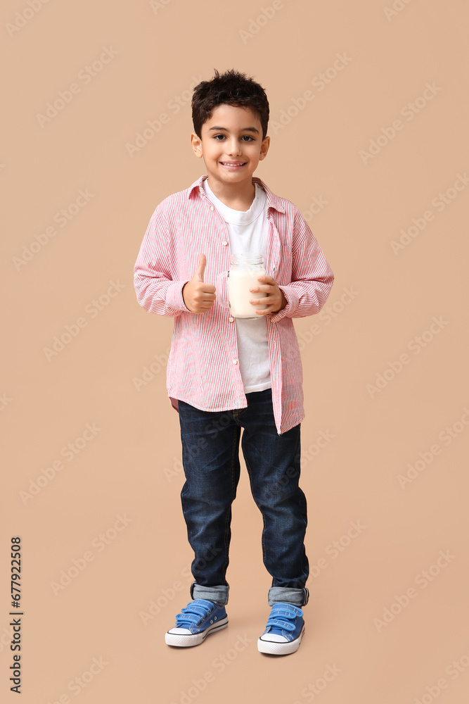 Cute little boy with mason jar of milk showing thumb-up on beige background