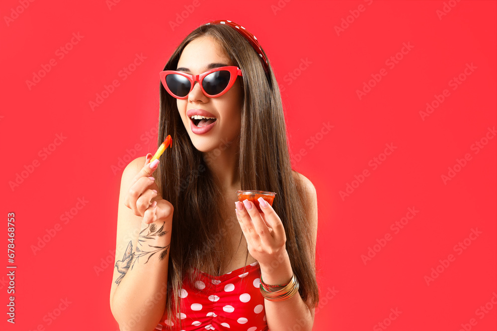 Beautiful young woman in sunglasses eating french fries with ketchup on red background