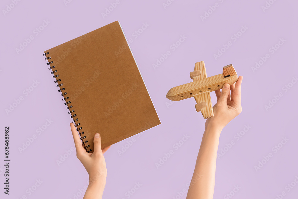 Female hands holding notebook and wooden airplane on lilac background