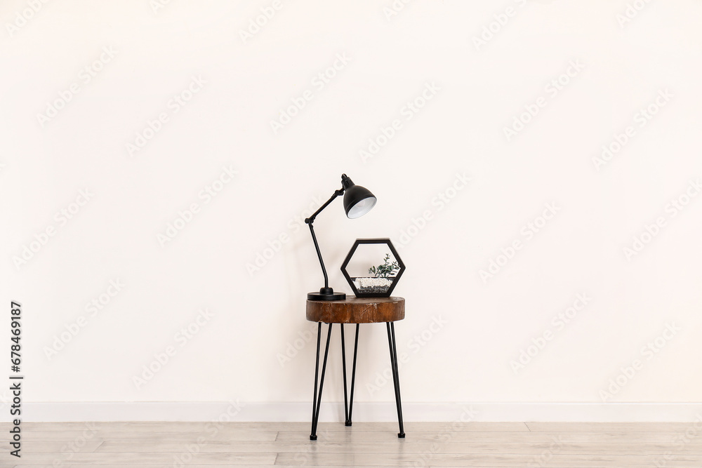 Desk lamp and florarium on small wooden table near white wall