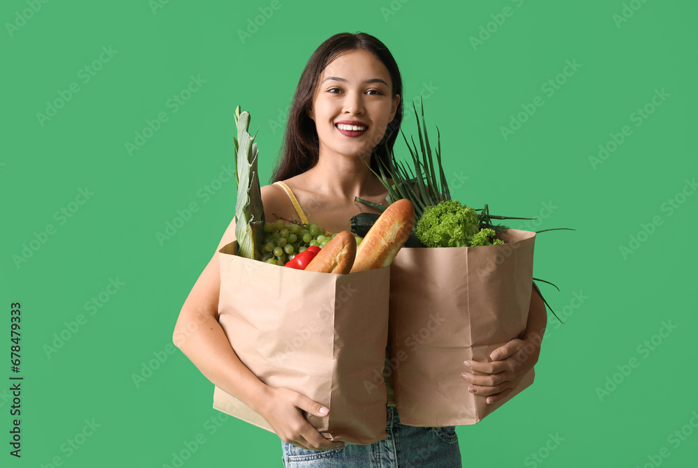 Young Asian woman with shopping bags full of fresh food on green background