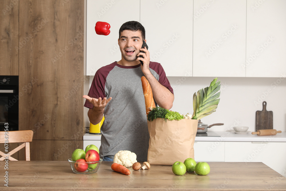 Young man with grocery bag talking by mobile phone at table in kitchen