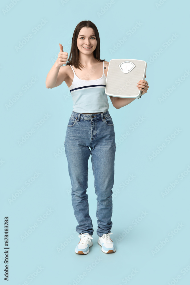 Beautiful young sporty woman with scales showing thumb-up gesture on blue background. Weight loss concept