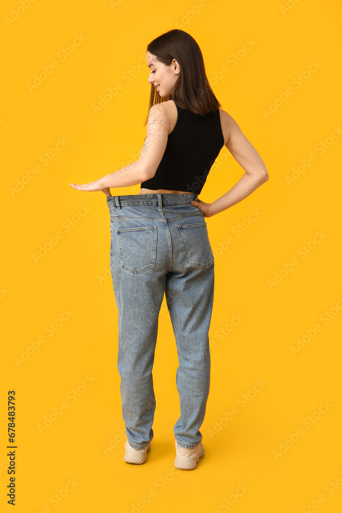 Beautiful young sporty woman in loose jeans on yellow background. Weight loss concept