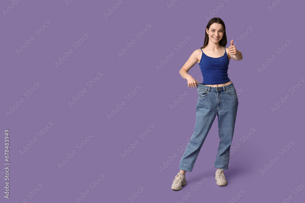 Beautiful young sporty woman in loose jeans showing thumb-up gesture on purple background. Weight loss concept