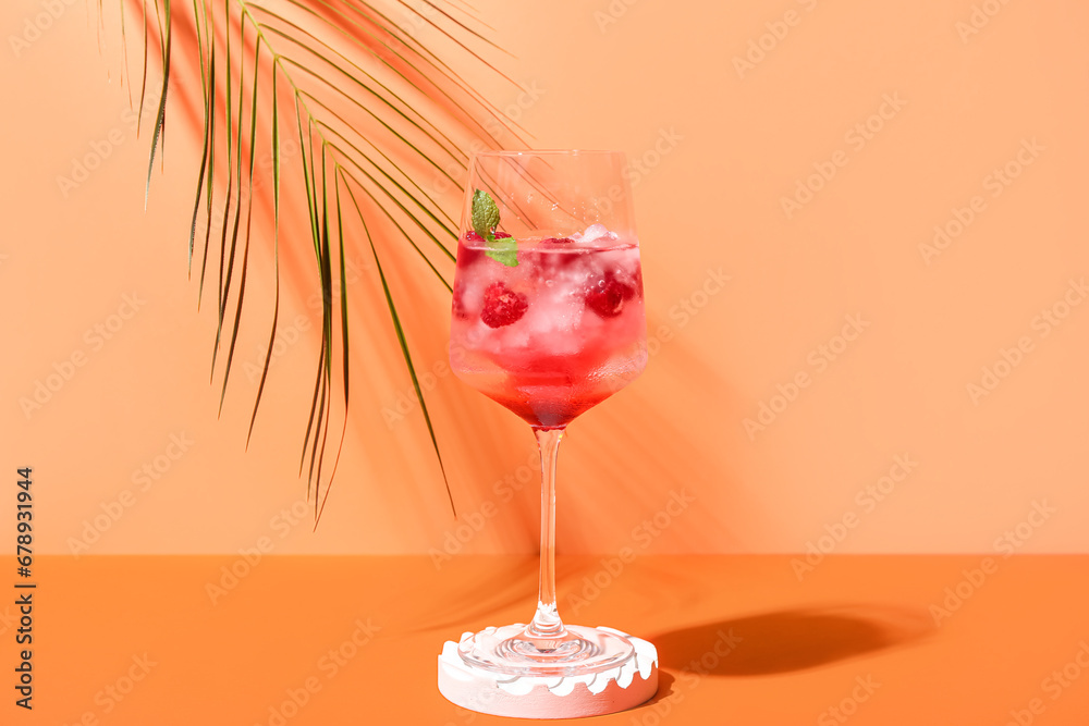 Decorative podium with glass of fresh raspberry mojito and palm leaf on orange background