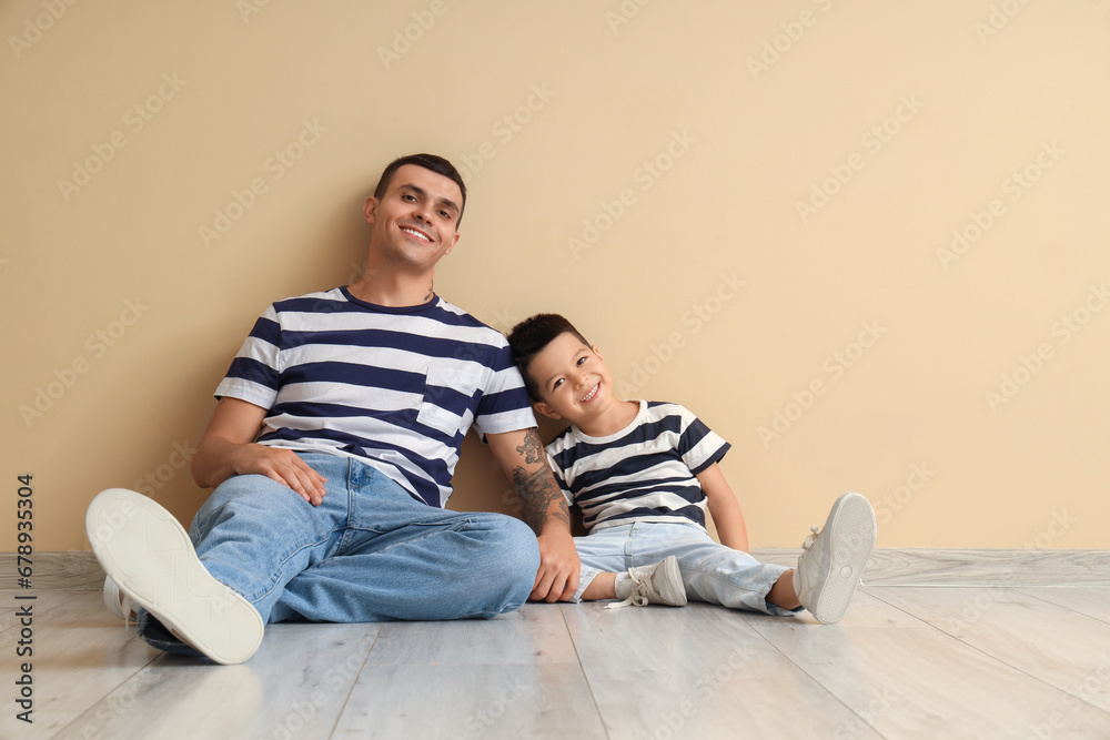 Young man with his little son sitting near beige wall