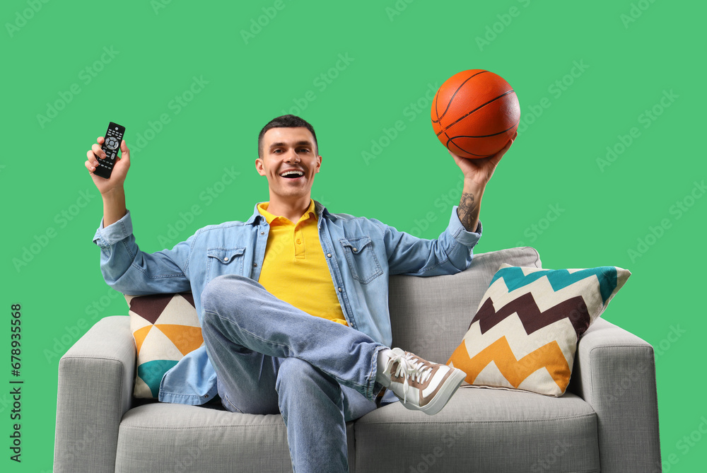 Young man with ball watching sport game on sofa against green background