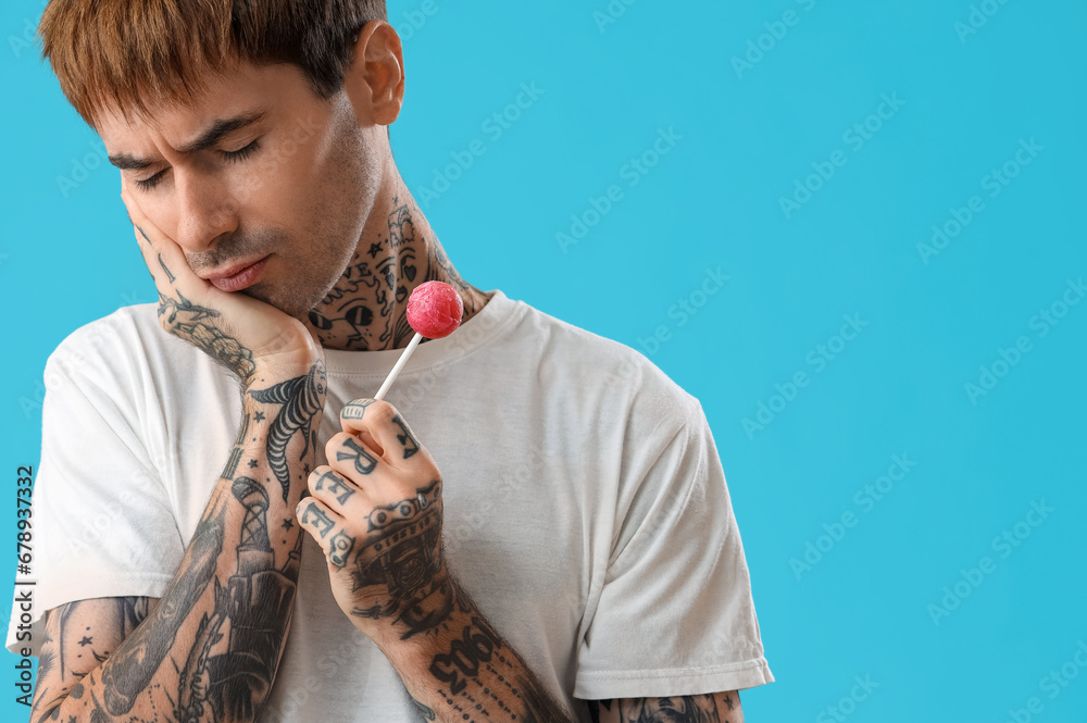 Young tattooed man with lollipop suffering from toothache on blue background, closeup