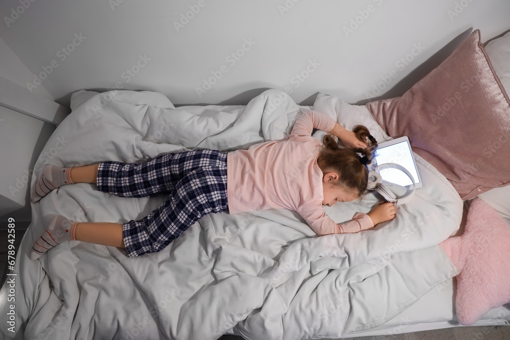 Little girl with headphones and tablet computer lying in bedroom at night, top view