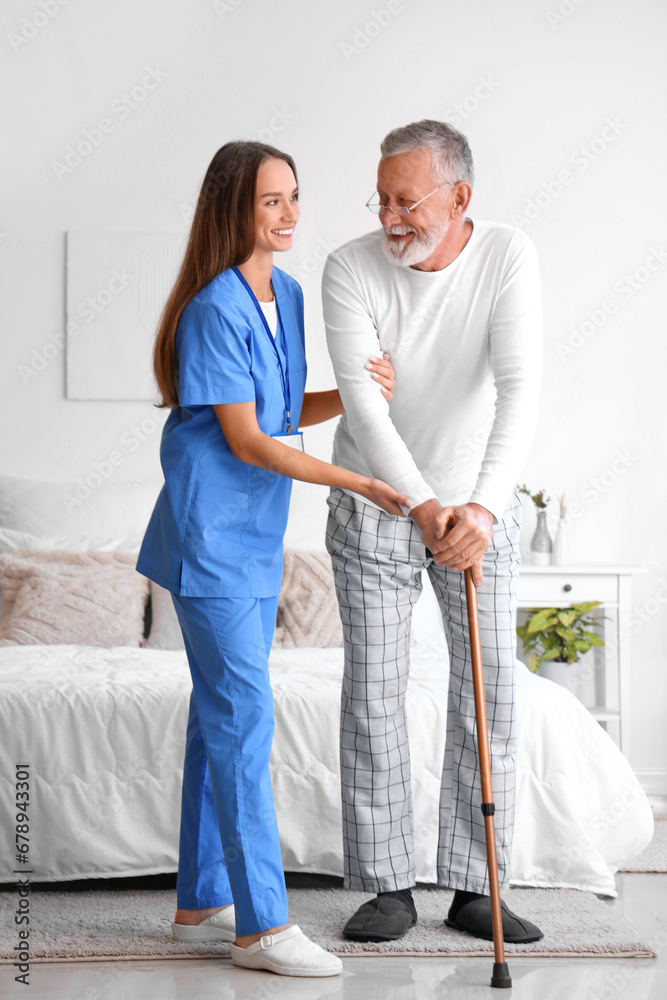 Female caregiver helping senior man with walking stick in bedroom